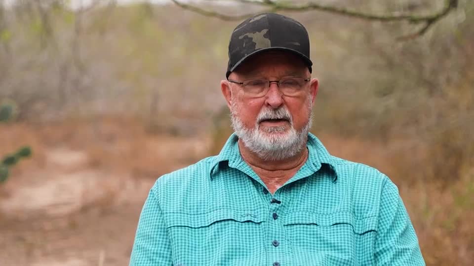 The man in the camo cap and glasses looks directly at the camera, wearing a blue plaid shirt. He's standing outdoors, with a blurred, earthy background.
