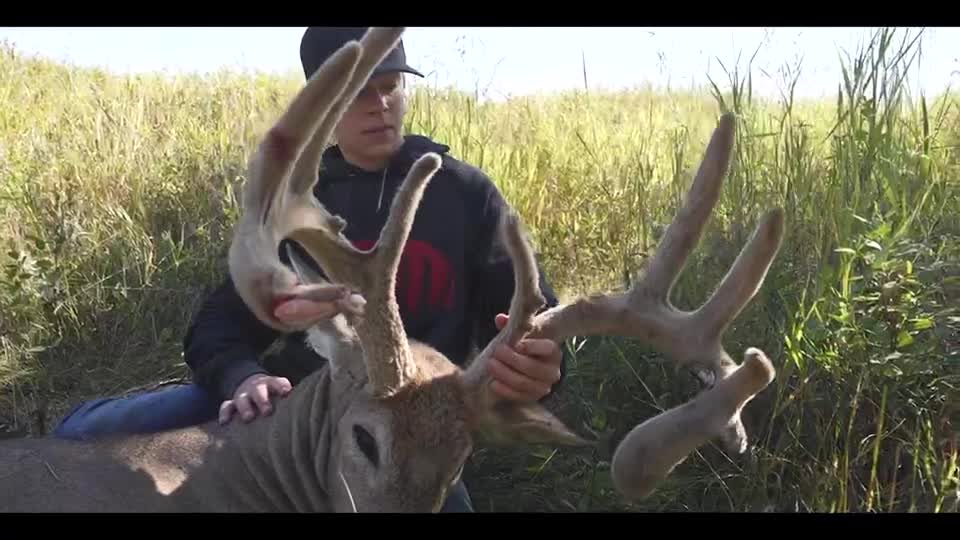 A young man kneels, holding the massive antlers of a buck. The animal lies on the ground in tall grass, its head resting against the man's leg.
