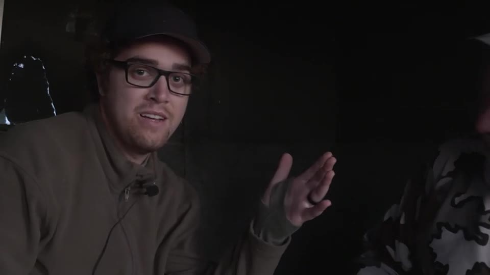 A man in a cap and glasses gestures with his right hand, speaking to someone out of frame. He's wearing a microphone and a green jacket, while the other person is clad in camouflage.
