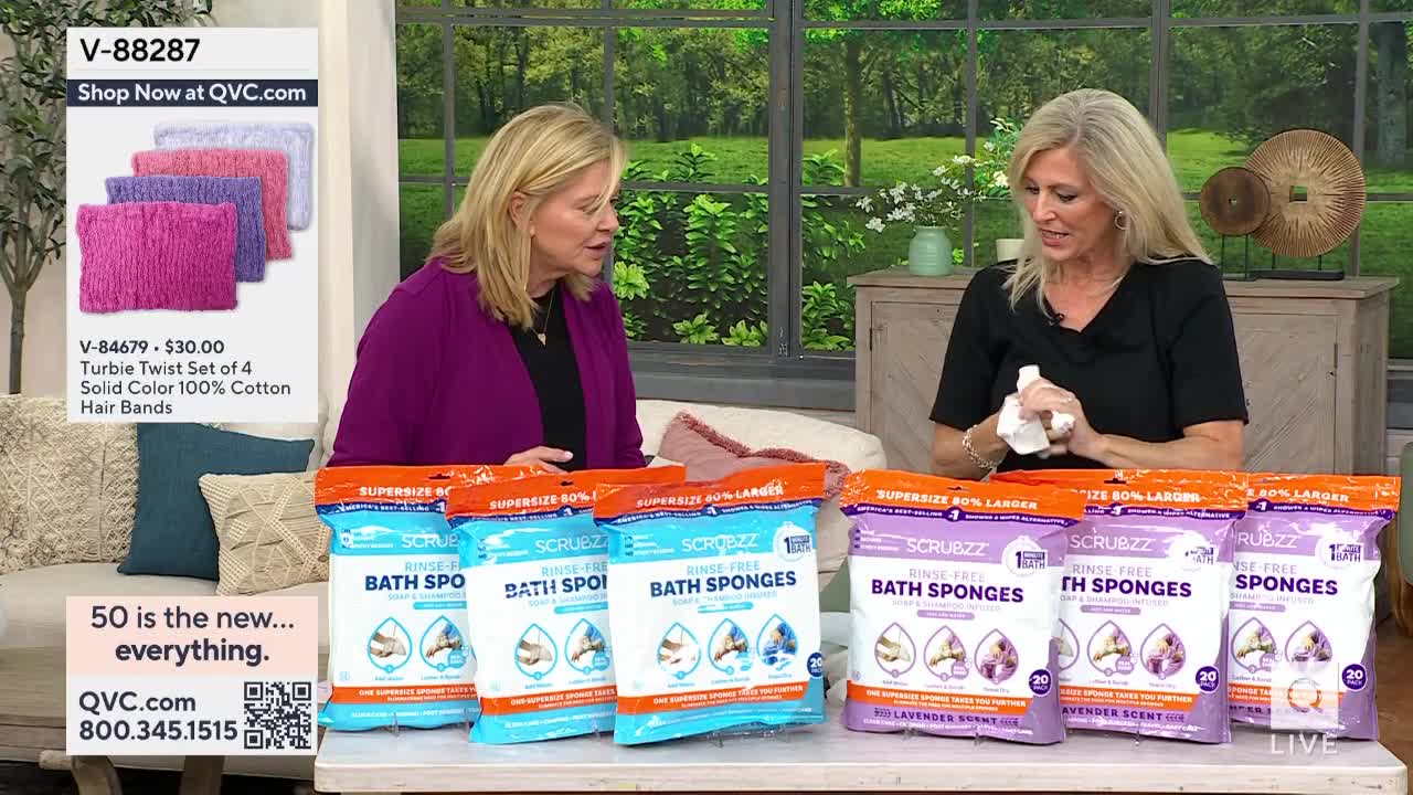 Two women are seated at a table, surrounded by packages of bath sponges, likely demonstrating them on a QVC broadcast. One holds a white cloth, while the other gestures towards a display of colorful hair bands.
