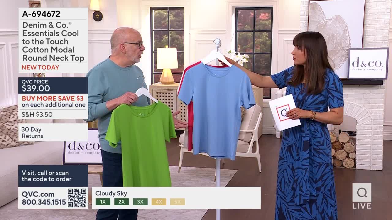 A man and a woman on a QVC set are holding up t-shirts on hangers. The woman gestures toward a blue shirt, while the man next to her displays a green one.
