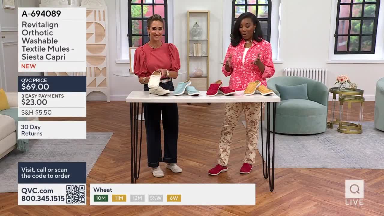 Two women stand on a set, presenting a variety of colorful shoes on a table. The QVC logo is visible, and the hosts are gesturing towards the footwear.
