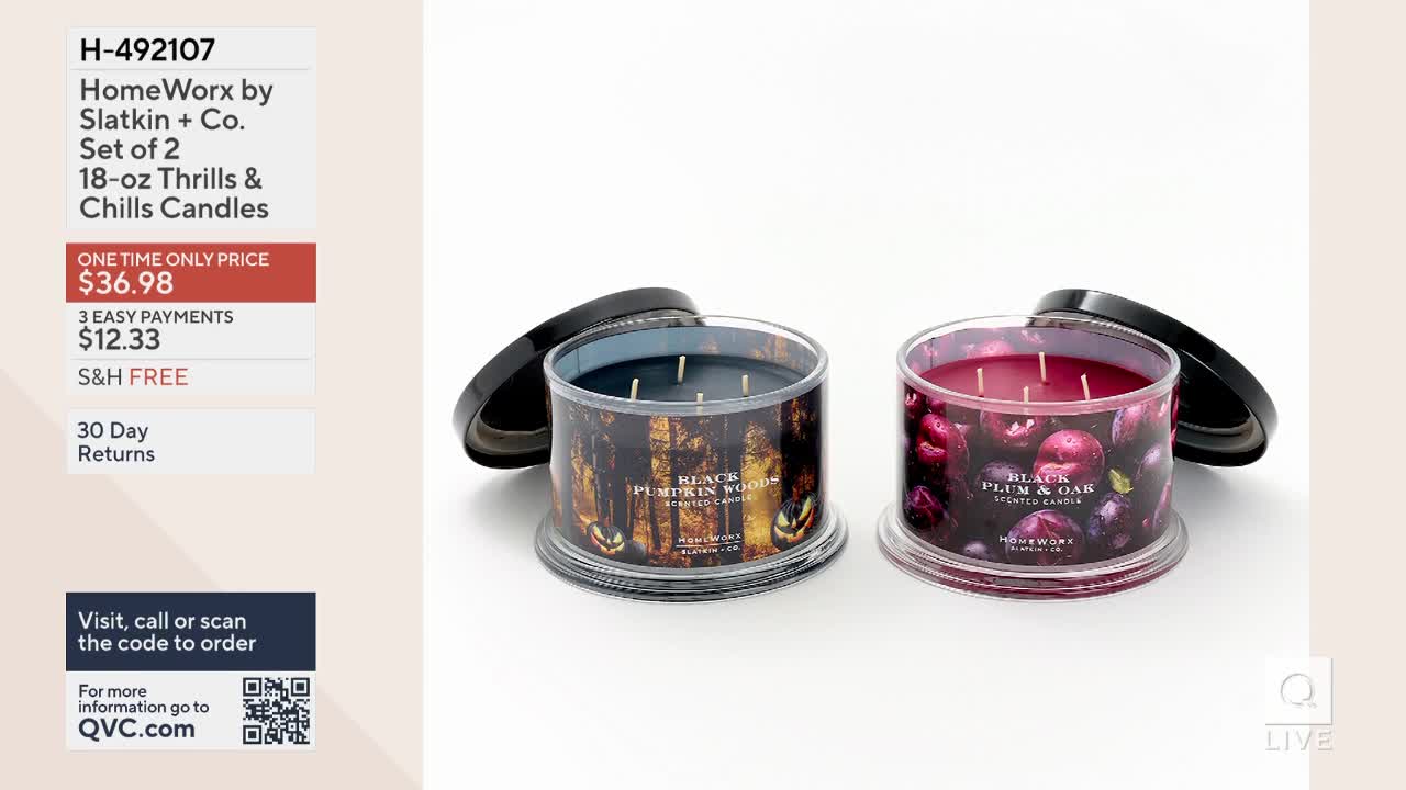 Two scented candles sit on a white surface, each with its lid slightly ajar. One is labeled "Black Pumpkin Woods," and the other "Black Plum & Oak," both advertised on QVC.
