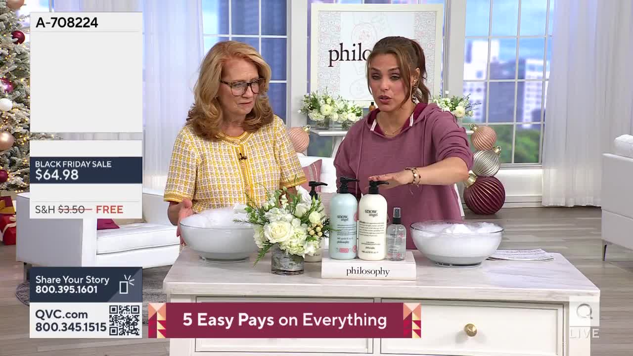 Two women stand behind a table displaying bath products, one gestures towards a bottle. Bowls of suds sit on either side of a small flower arrangement, and a screen in the corner advertises a Black Friday sale on QVC.
