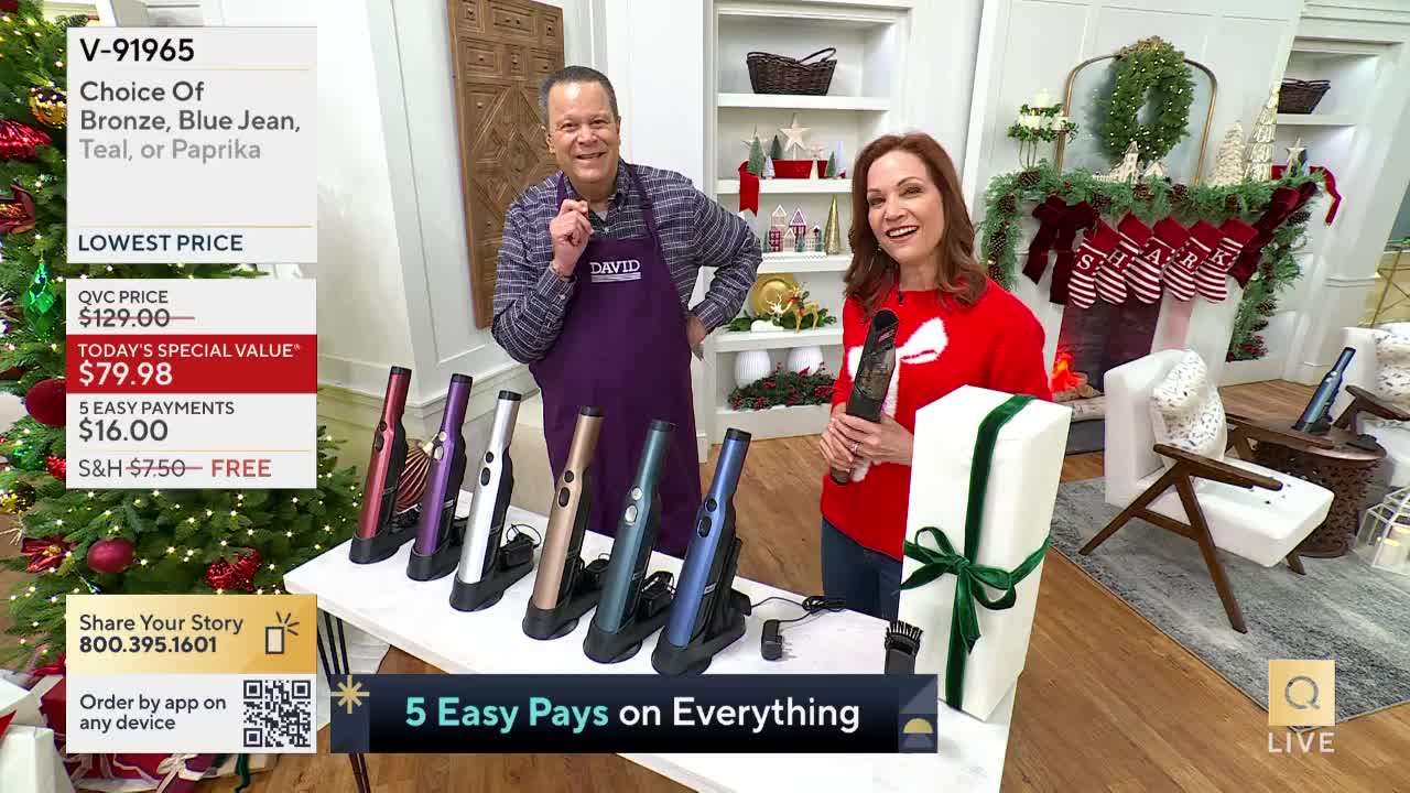 A man and woman stand behind a table displaying several cordless hair stylers, the woman holding one up. The QVC hosts are smiling, and the set is decorated for the holidays.
