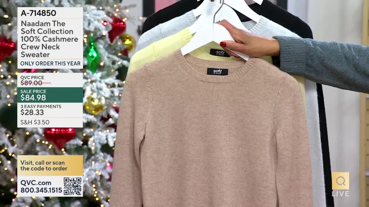 A hand, wearing a gray sweater, is holding up a light brown cashmere crew neck sweater on a hanger. Behind it, other sweaters in different colors hang on a rack, all on display for a live QVC segment.
