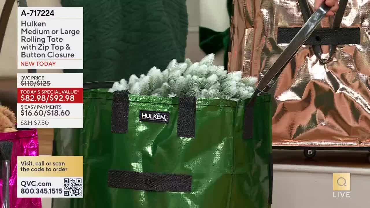A green tote bag, filled with white, fluffy objects, is being demonstrated on a QVC set. The presenter is holding the handle of a similar, metallic bag, perhaps showing how it rolls.
