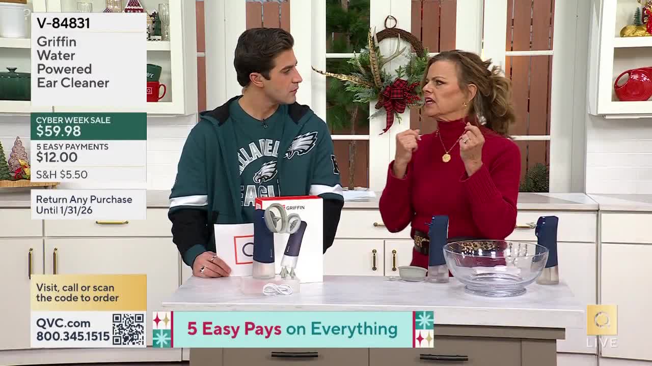 The woman in the red turtleneck gestures with her hands, possibly demonstrating something, as a man in an Eagles sweatshirt looks on. On the countertop, several blue and white devices sit next to a clear bowl, likely for a product demonstration on QVC.
