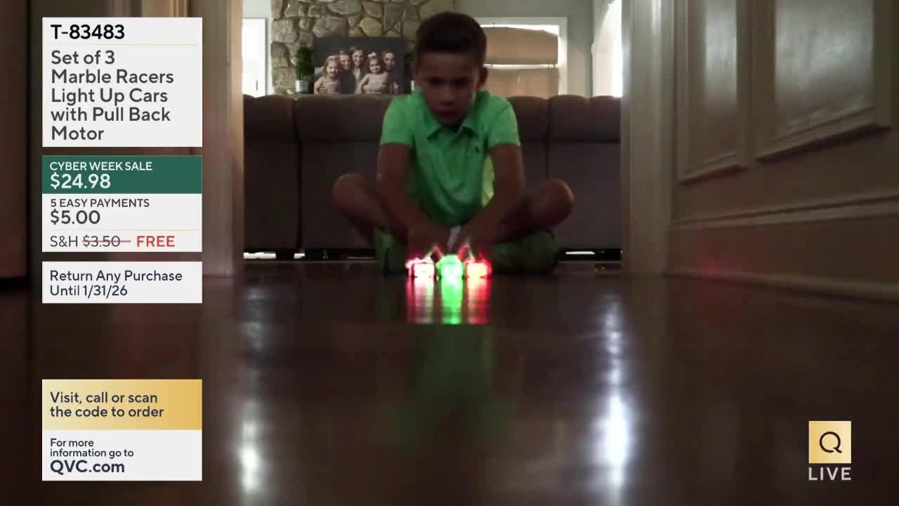 A young boy sits cross-legged, focused on three small, glowing toy cars. The cars, lit with red and green lights, are lined up on a shiny wooden floor.
