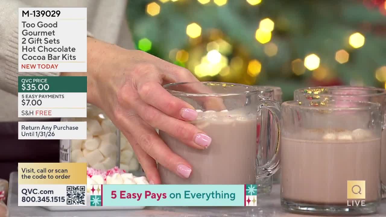 A hand with pink nails gently holds a clear mug filled with hot chocolate, topped with marshmallows. The scene is part of a QVC presentation, with a product description visible on the screen.
