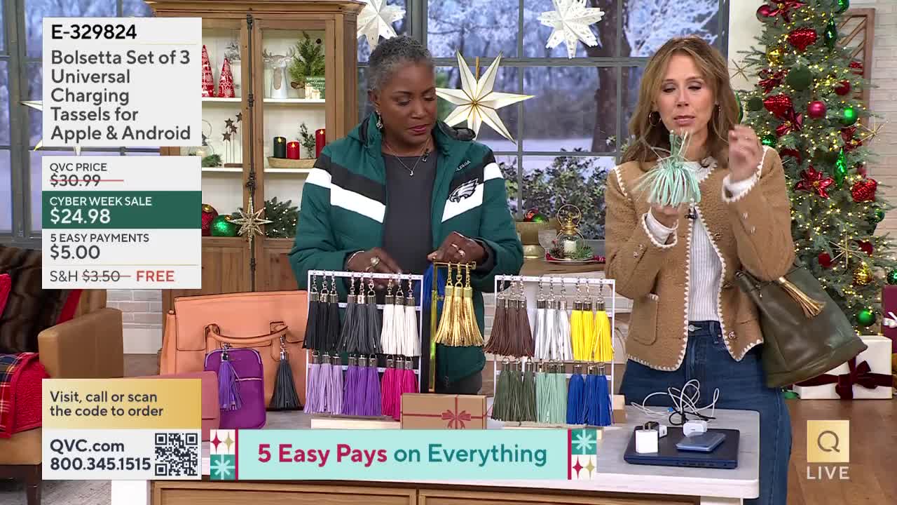 A woman in a blazer holds up a tassel, demonstrating its use with a charging cable and phone. Another woman, wearing an Eagles jacket, gestures towards a display of colorful tassels on a rack, likely for sale on QVC.
