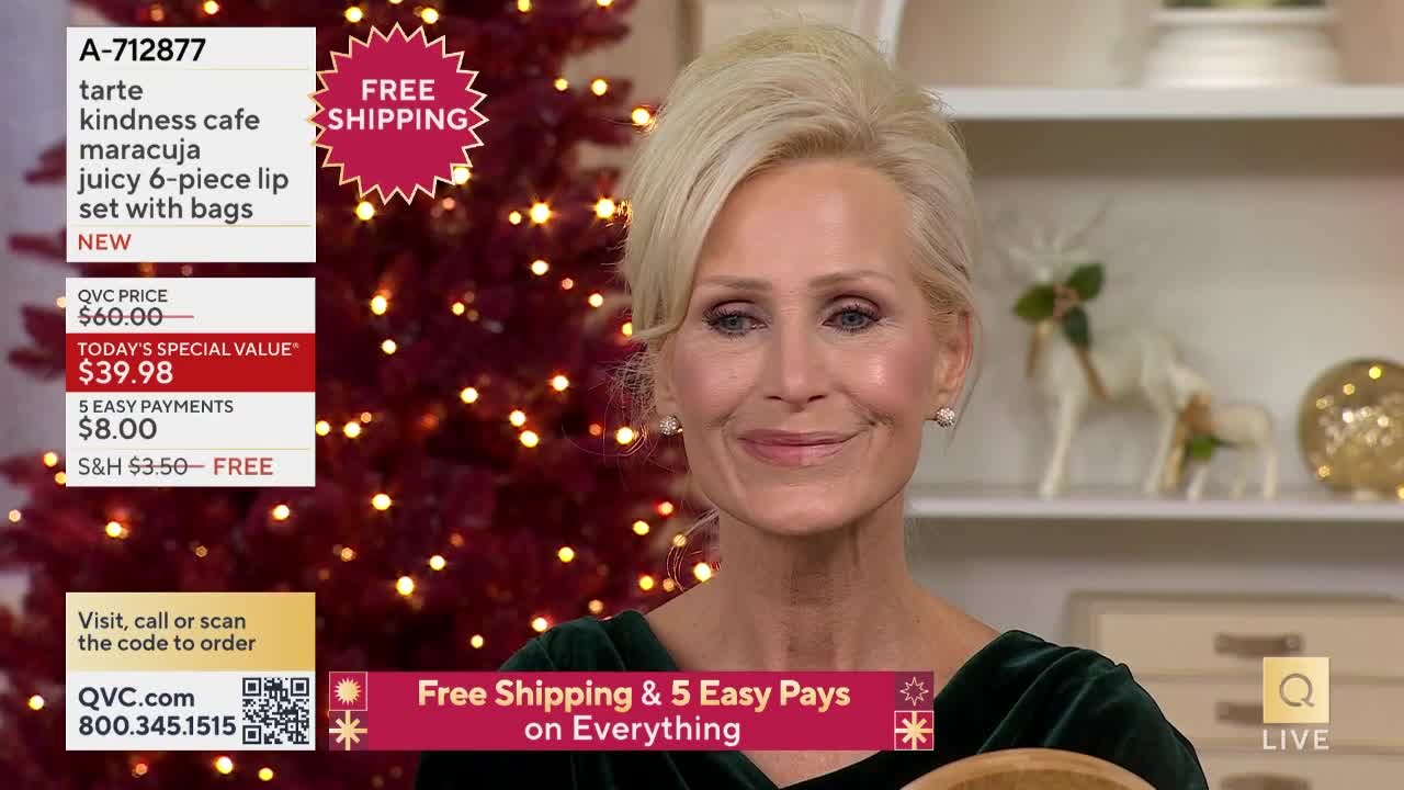 A woman with blonde hair smiles at the camera, holding something out of frame. Behind her, a Christmas tree glows with tiny lights, and the QVC logo is visible on the screen.
