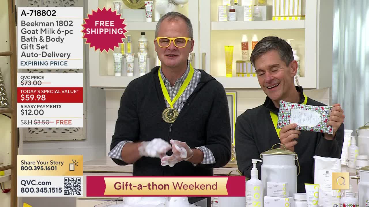One man, covered in suds, gestures with his hands while another holds up a package, both smiling at the camera on a QVC set. A display of bath products and promotional information surrounds them.
