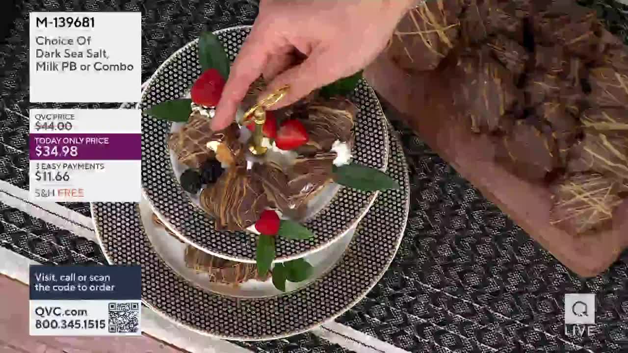 A hand with a gold ring carefully places a small golden ornament on a tiered dessert. This confection, adorned with strawberries and dark chocolate swirls, sits on a patterned plate.