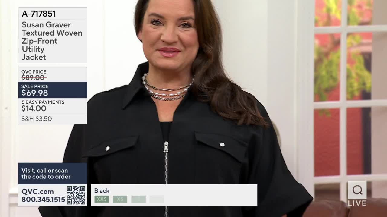 A woman stands in a black utility jacket, a QVC logo visible on the screen. The jacket features a silver zipper and two chest pockets.