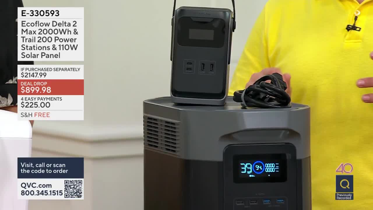 A presenter in a bright yellow shirt gestures towards a large, dark gray power station. A smaller, black portable power unit sits on top of it, with a coiled black cable resting beside it.