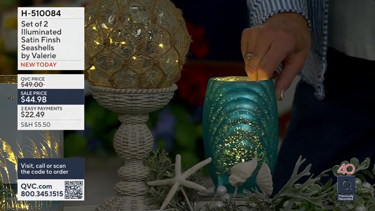 A hand reaches into a teal glass hurricane, its surface textured with raised ridges. Tiny lights glow within the hurricane, mirroring the illumination from a seashell decoration on a pedestal to its left.
