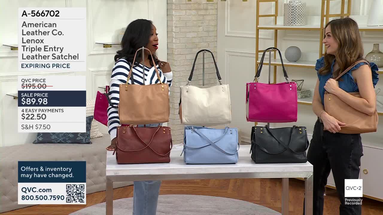 Two women stand in front of a table displaying several leather satchels in various colors. One woman holds a tan bag, while the other gestures towards the assortment of bags on the table.
