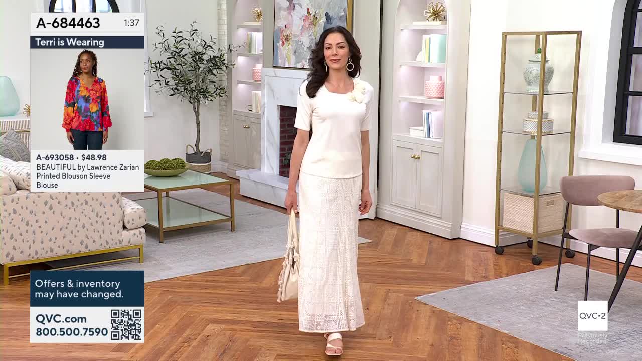 A woman in a cream-colored outfit strides across the set, holding a matching handbag. A screen shows a model wearing a colorful blouse, and the QVC2 logo is visible in the corner.
