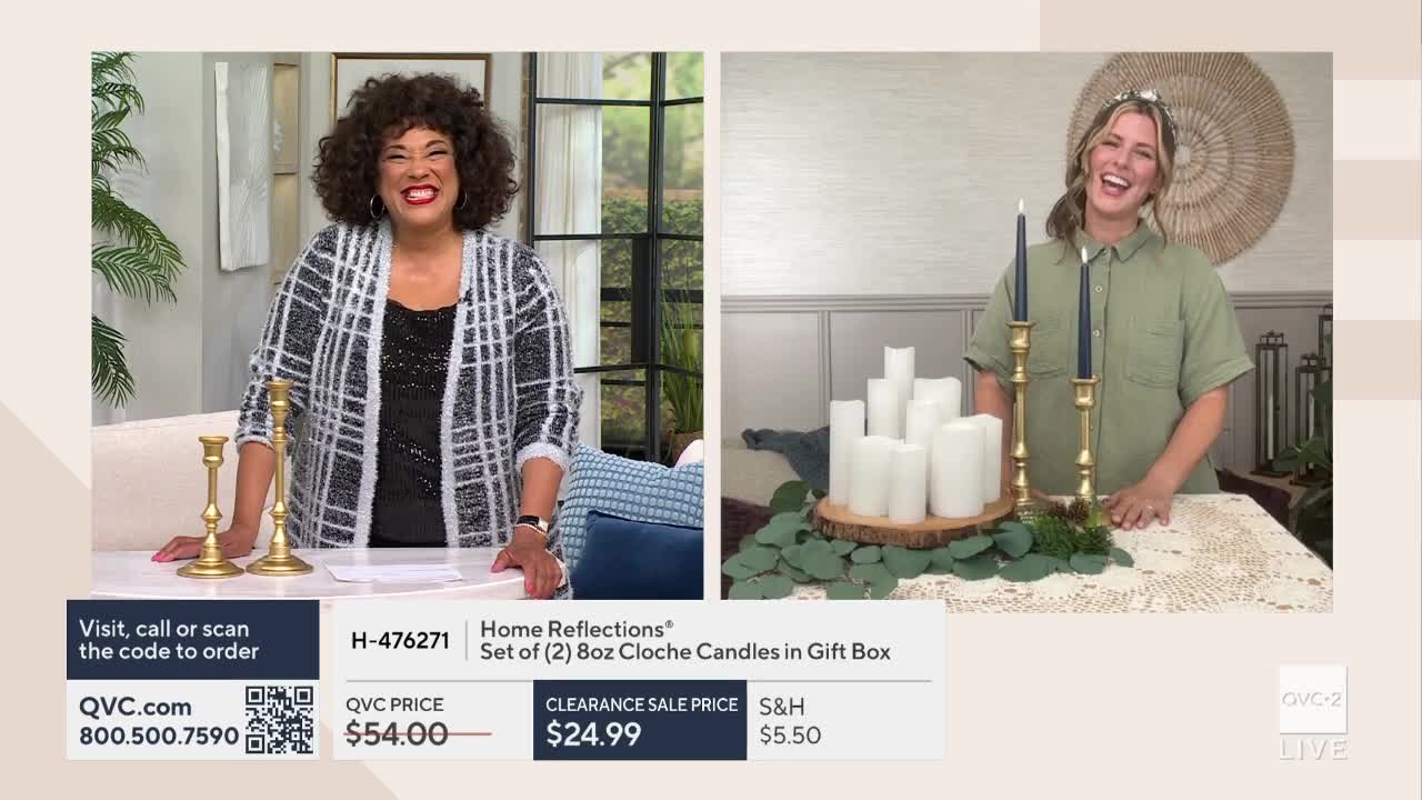 Two women on a QVC2 set are enthusiastically presenting candles. One woman stands near gold candlesticks, while the other showcases a display of white candles on a wooden base.
