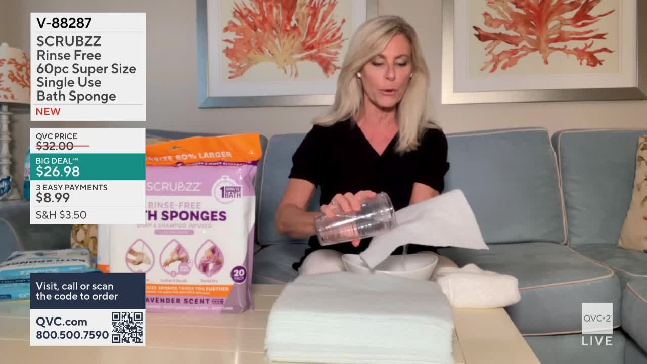 On QVC2, a woman in a black top is demonstrating the bath sponges. She's pouring liquid from a clear cup onto a stack of white, absorbent pads.
