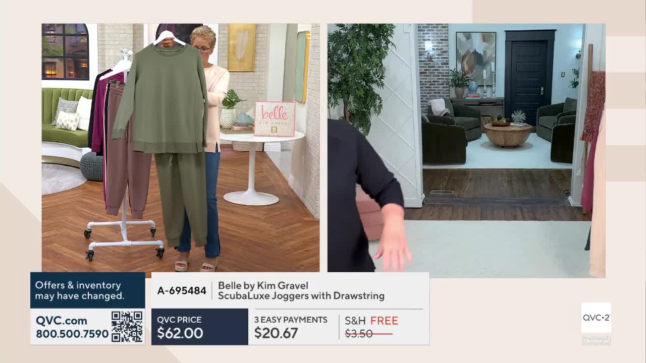 A woman in jeans holds up a green sweatsuit in front of a rolling rack displaying other colors. The QVC2 set has a living room visible in the background.
