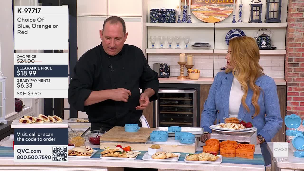 A man in a black shirt gestures toward a table laden with pastries and blue cookie cutters. Across from him, a woman in a blue blazer looks on, a plate of food in front of her.
