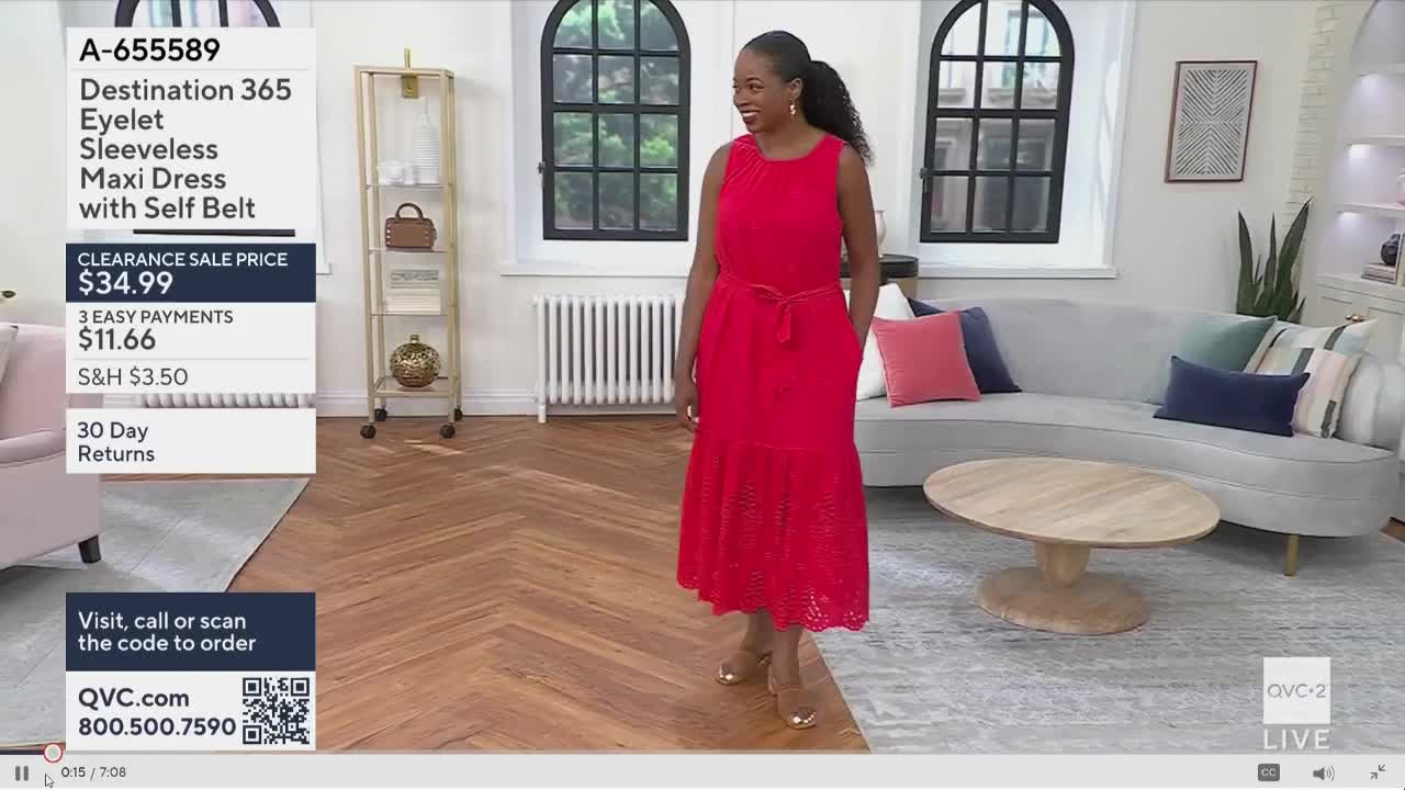 A woman in a bright red dress is walking toward the camera on a QVC2 set. She smiles, modeling the dress, which is on sale for $34.99.
