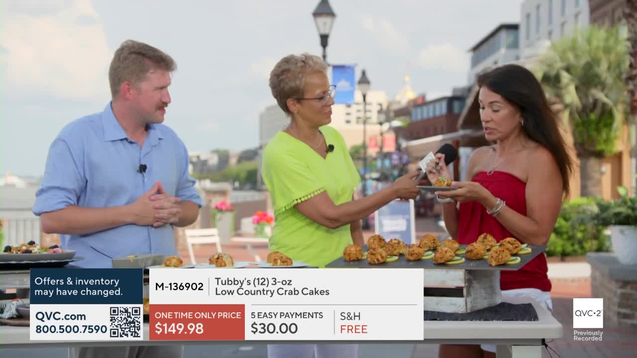 A woman in a red top holds a crab cake, ready to eat it while speaking into a microphone. Two other people stand nearby, and a display of crab cakes sits on a table in front of them.
