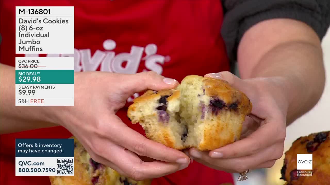 A woman's hands are pulling apart a blueberry muffin, revealing its soft, yellow interior. The QVC2 logo is visible in the corner.
