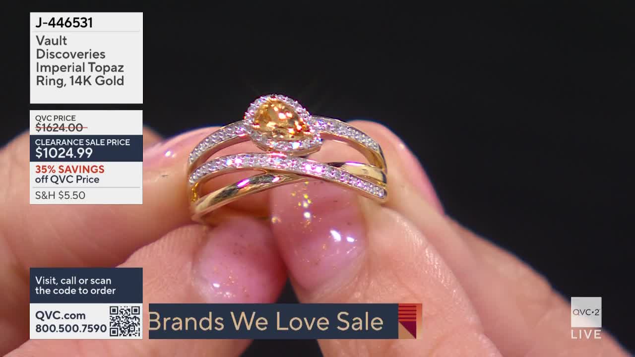 A hand, with painted nails, holds a gold ring with a large, light orange stone. The ring's price and details are displayed on a screen from QVC2.
