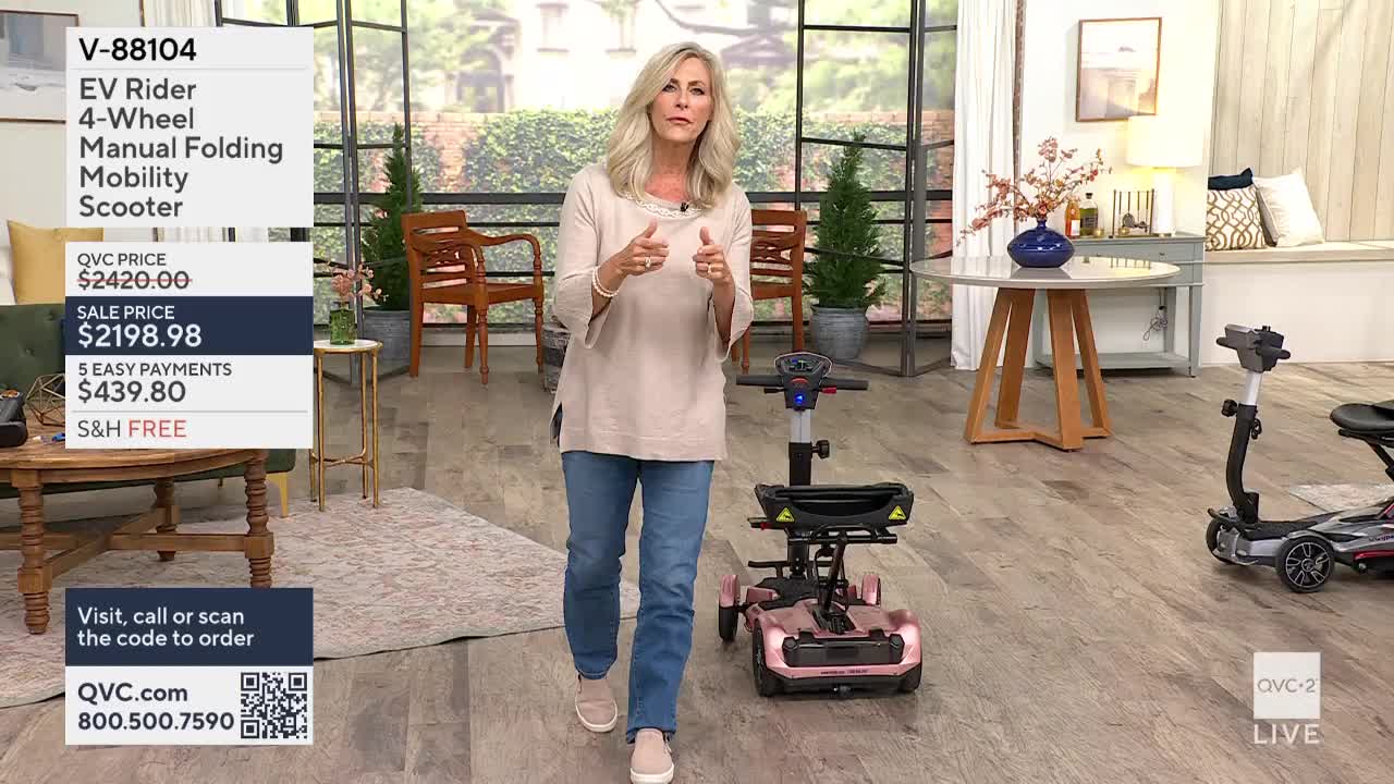 A woman in jeans and a light-colored top gestures towards a pink mobility scooter. The QVC2 set features a few other scooters and some decorative plants.
