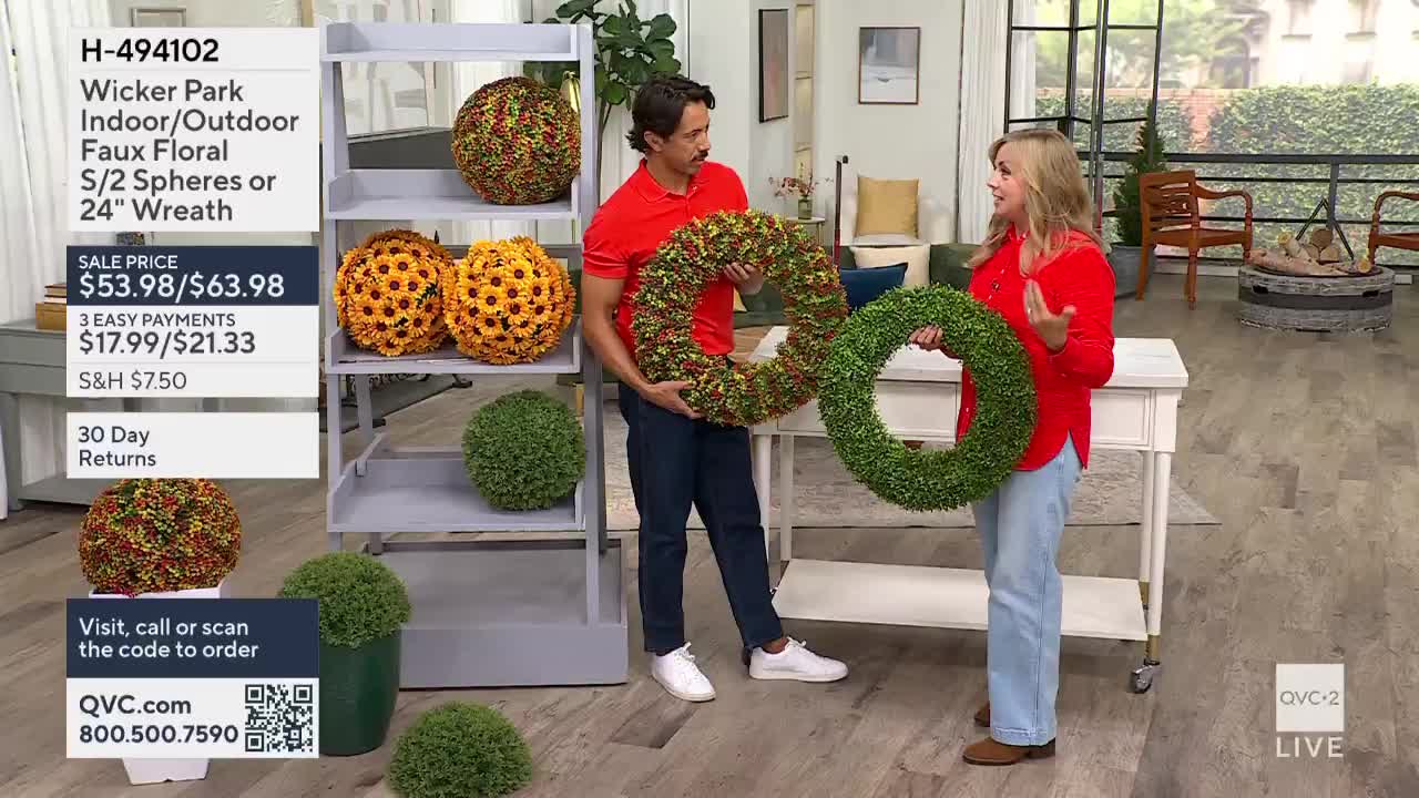 A man and a woman, both in red shirts, hold up large, artificial wreaths. Behind them, a shelf displays more faux floral spheres and wreaths, all ready for sale on QVC2.
