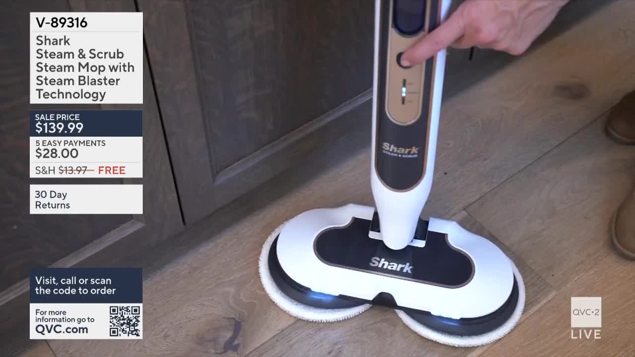 A hand presses a button on a Shark steam mop, activating it on the wooden floor. The QVC2 graphic shows the mop's price and ordering information.
