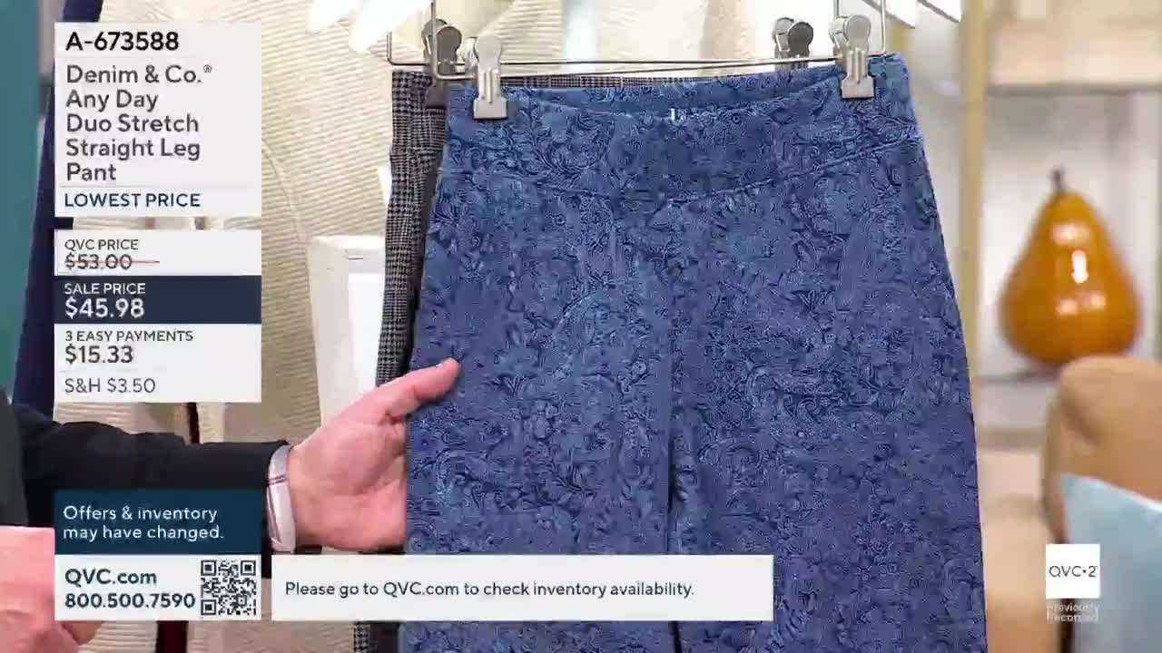 A man's hand holds up a pair of blue patterned pants, showing them off on QVC2. The camera focuses on the pants, while a price tag and website information are visible in the corner.
