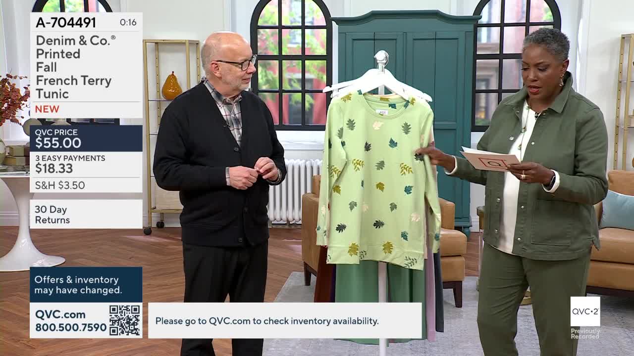 A woman in a green jacket holds a light green tunic with leaf patterns on a hanger. A man in a cardigan stands beside her as she reads from a card, all while on QVC2.
