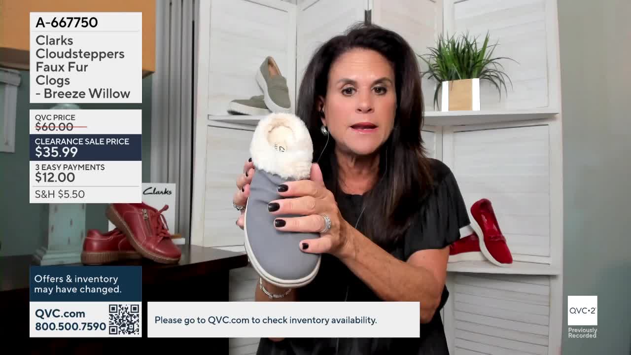 A woman on QVC2 holds up a gray clog with a fluffy, cream-colored lining. Behind her, a pair of red shoes and another pair of gray ones sit on shelves.
