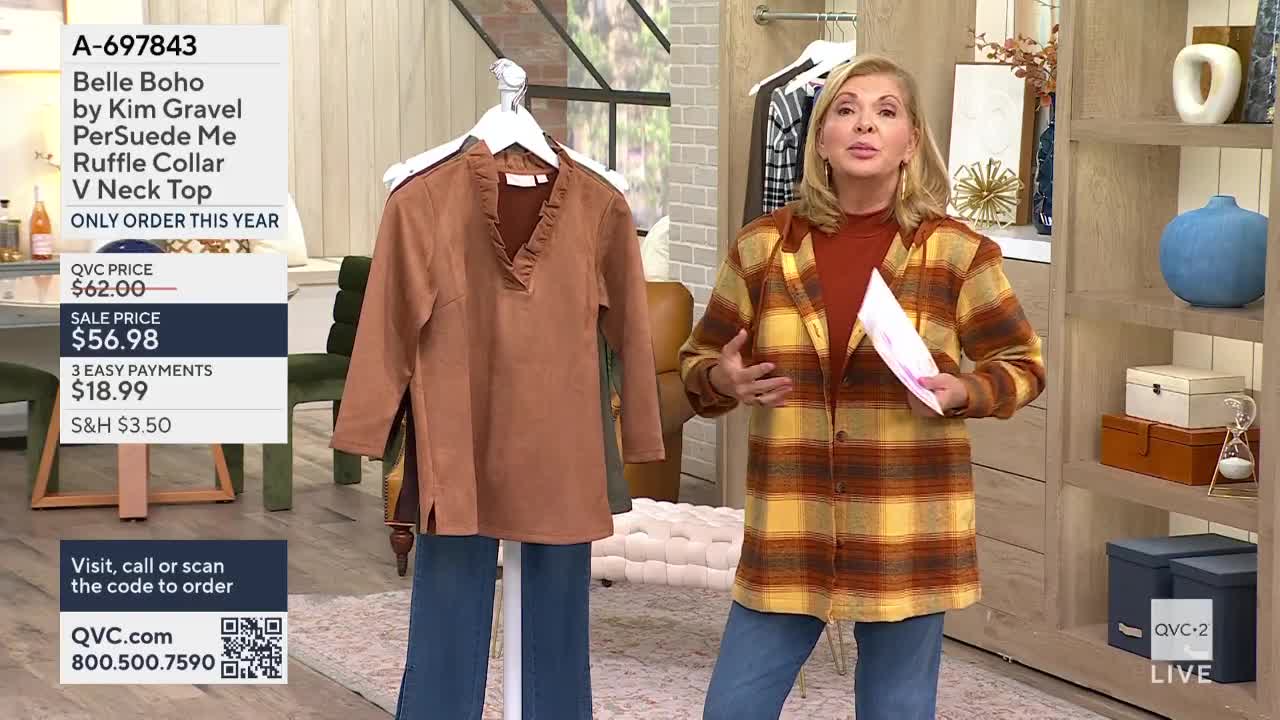 A woman on QVC2 gestures with a paper in her hand while describing a brown top hanging on a rack. Beside it, a pair of jeans are displayed, and the presenter wears a plaid jacket over a rust-colored shirt.
