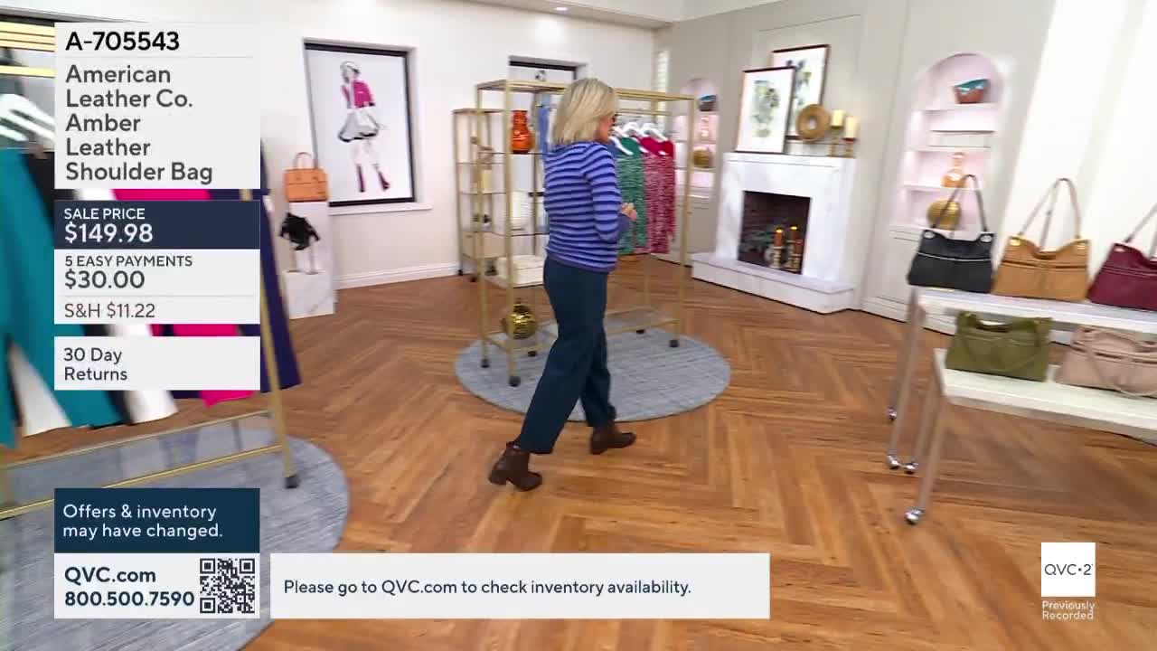 A woman in a blue striped shirt and dark pants walks across the wooden floor. The scene is from a QVC2 broadcast, with product information displayed on the left.

