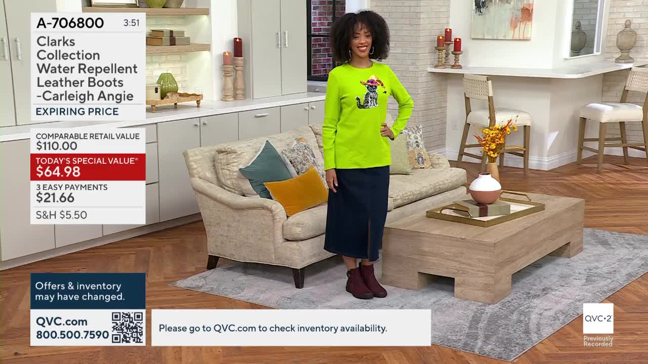 A woman stands in a studio, wearing a bright green shirt and dark skirt, modeling burgundy boots. The QVC2 logo is visible in the corner, with a product description displayed on a screen.
