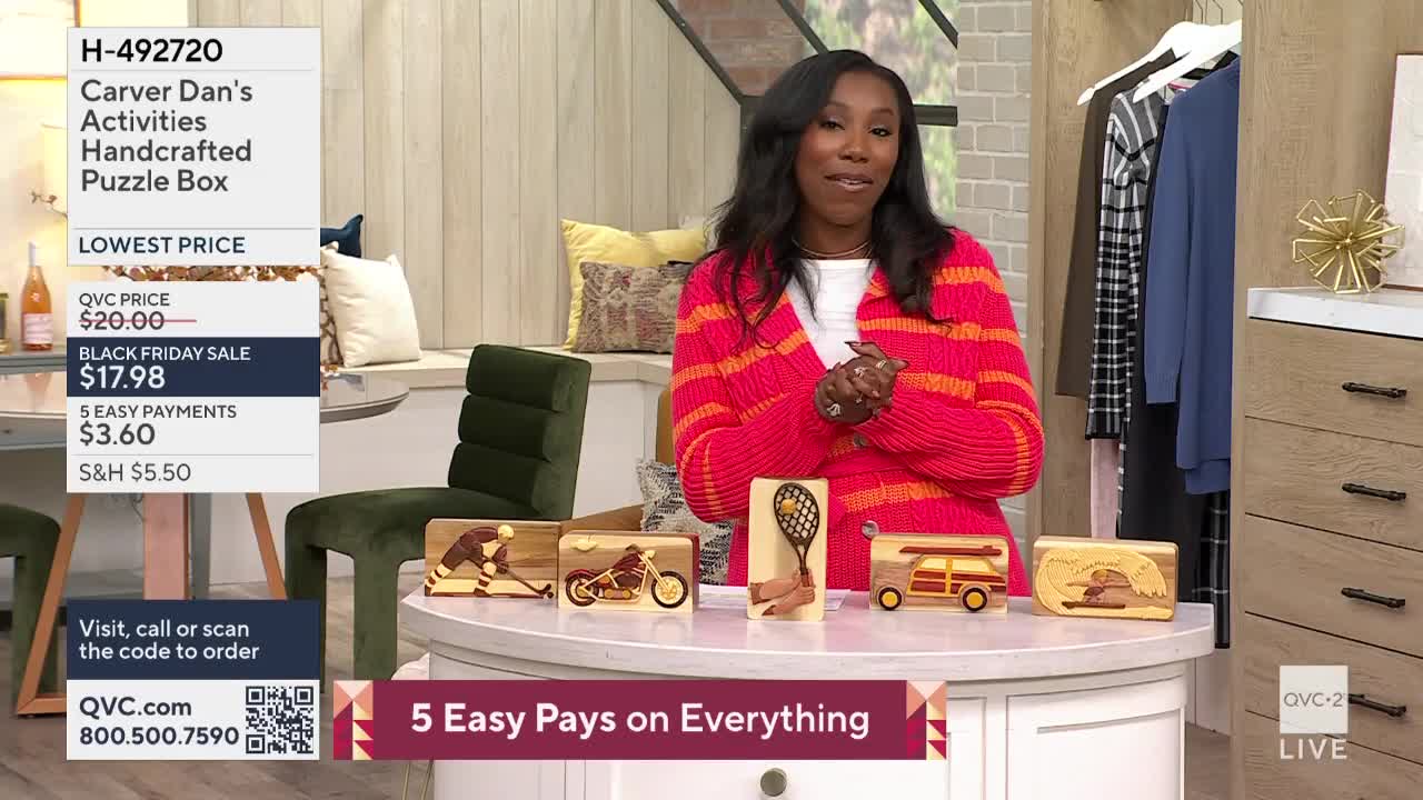 A woman in a bright, striped sweater stands behind a table displaying wooden puzzle boxes. The boxes are arranged in a row, each depicting a different activity, and a QVC2 graphic is visible in the corner.
