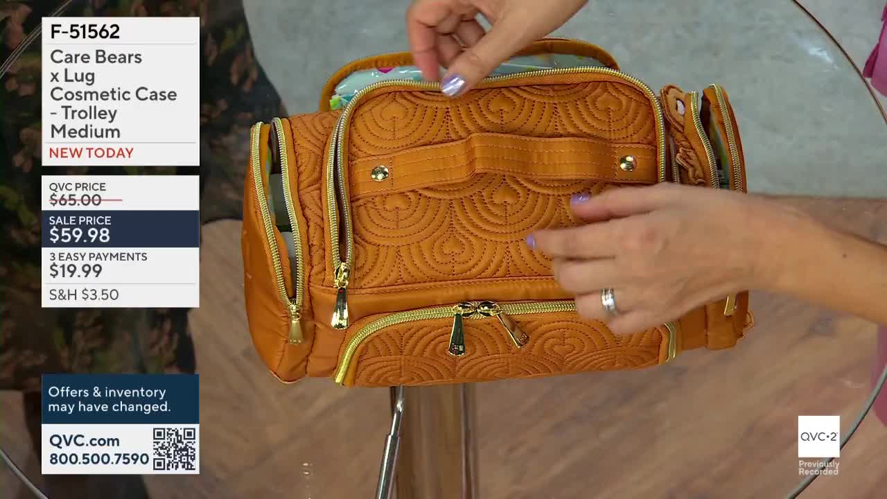 A woman's hands are unzipping a quilted orange cosmetic case on QVC2. The bag has gold zippers and a handle, and the presenter is showing off its compartments.
