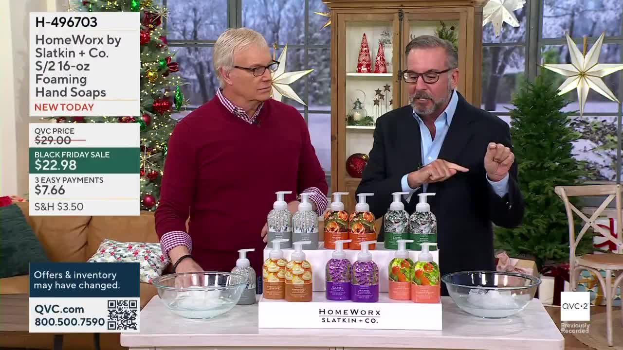 Two men stand behind a table displaying various bottles of soap. One gestures with his hand while the other looks on, both framed by holiday decorations, all on QVC2.
