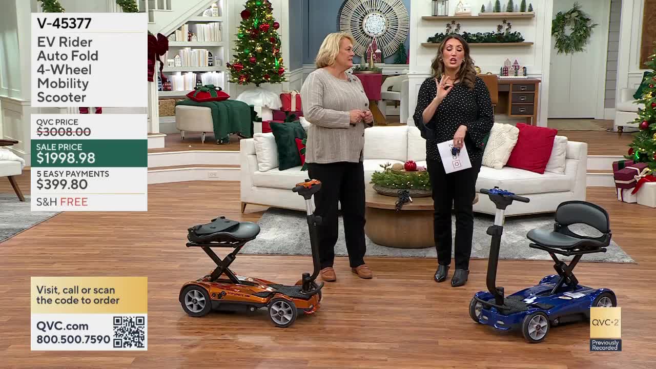 Two women stand in a decorated living room, gesturing towards two mobility scooters. The orange and blue scooters are displayed on a hardwood floor, with the QVC-2 logo visible in the corner.
