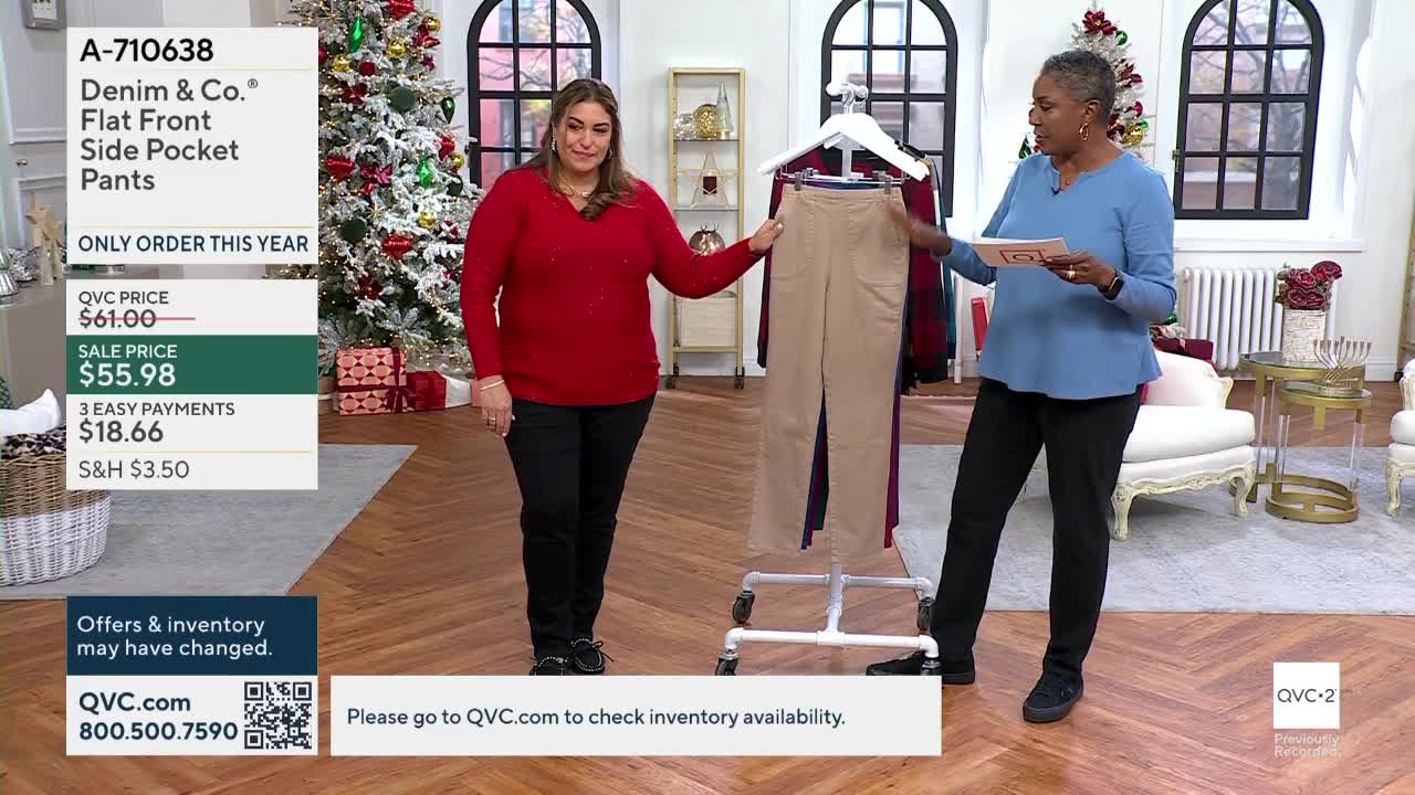 Two women stand on a set, one in a red sweater gesturing at a pair of tan pants on a rolling rack. The other woman, dressed in blue, holds a small card and points towards the pants as well.
