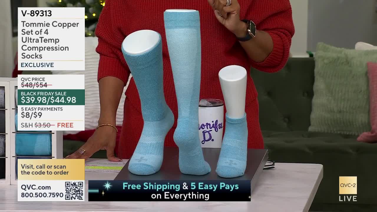 On a table in a QVC2 studio, four compression socks are displayed on white mannequins. A host in a red sweater gestures towards the socks, highlighting the Black Friday sale.

