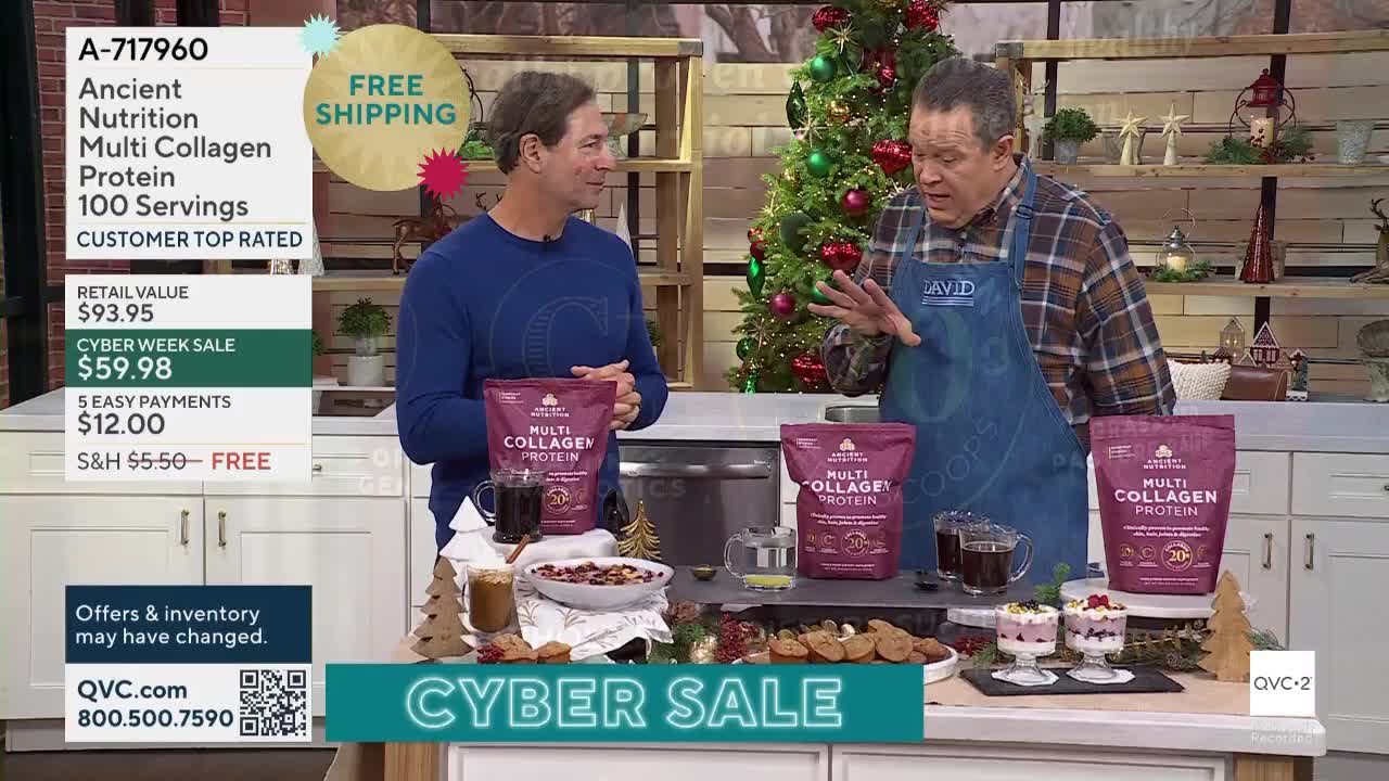 Two men stand in a kitchen setting, holding bags of protein powder as they talk. The man in the apron gestures with his hands, while the other man smiles and holds a bag of the product.
