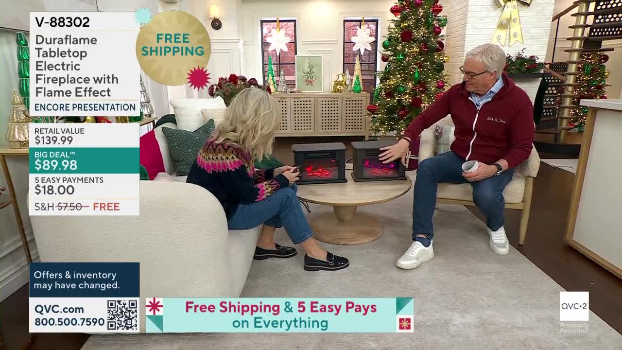 The woman is looking at one of the small electric fireplaces on the table, while the man points to the other. A Christmas tree and other festive decorations are visible in the background, as this is a presentation on QVC2.
