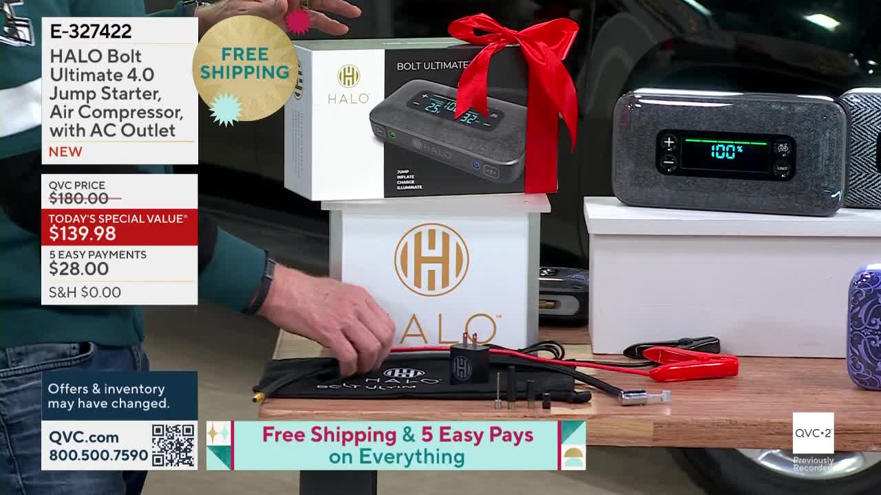 A man is holding a black case with cables and clamps, likely part of the Halo Bolt Ultimate 4.0 jump starter being promoted on QVC2. The product's box, topped with a red bow, sits on a white pedestal.
