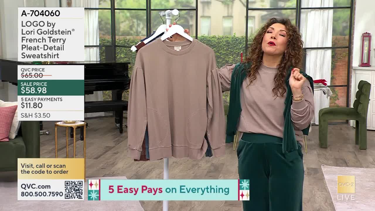 A woman in green velvet gestures toward a taupe sweatshirt hanging on a rack. The QVC2 host is standing in a studio, promoting the item.
